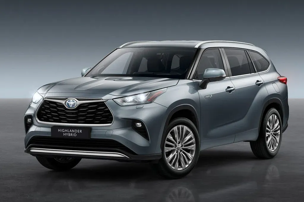 Toyota Highlander-style three-row SUV driving on a suburban road with an electric vehicle charging station in the background