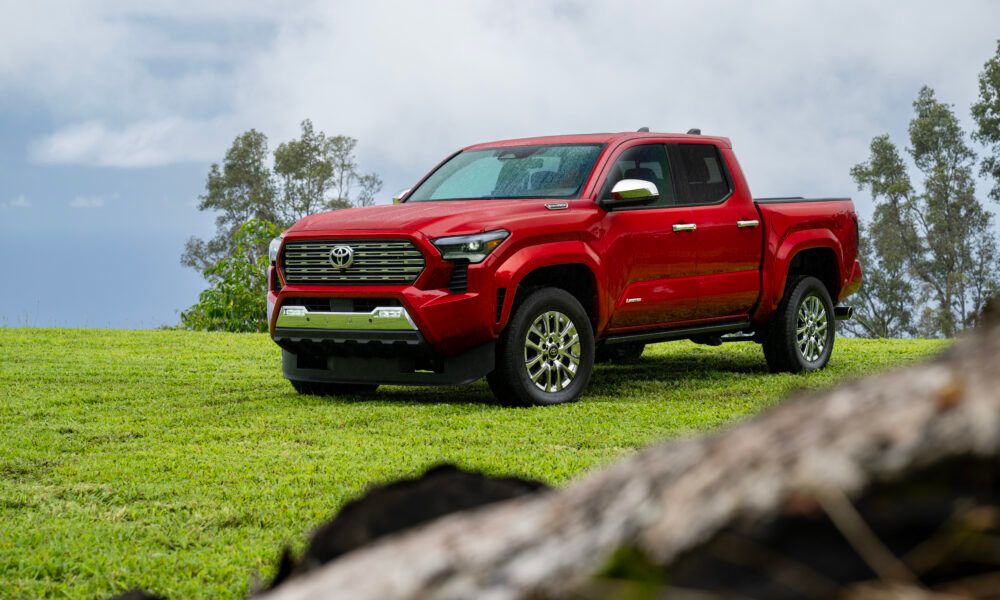 A current-generation Toyota Tacoma driving on a dirt road with a factory-style industrial backdrop in the distance